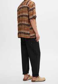 Kortærmet, flerfarvet mønstrede skjorte med vandrette striber, båret med sorte løst siddende bukser og lysebrune slip-on sko.