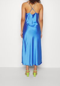 Satin blue maxi dress with spaghetti straps and crisscross back design. Paired with green block-heel sandals, showcasing a smooth texture.