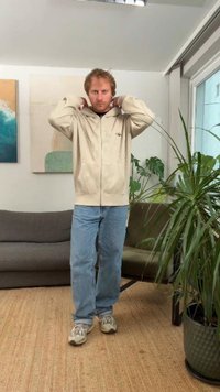 Beige-vetoketjullinen huppari, jossa logo, mustan paidan päällä, yhdistetty vaaleansinisiin farkkuihin ja harmaisiin lenkkareihin, seisoo huoneessa, jossa on kasveja.
