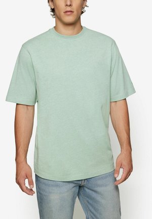 T-shirt vert clair à manches courtes en tissu doux et texturé ; présente un col rond et une coupe décontractée, associé à un jean en denim bleu.