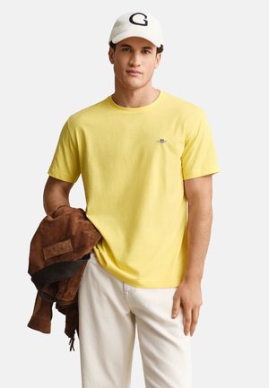 Uomo con t-shirt gialla e cappellino bianco che tiene una giacca marrone, in piedi davanti a uno sfondo chiaro uniforme.