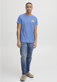 Blå kortermet T-skjorte med rund hals, med et lite palmetre-trykk på venstre bryst. Brukt med blå denimjeans.