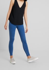 Femme portant un jean skinny bleu, un haut noir sans manches à col en V, et des baskets compensées beige clair, debout sur un fond gris uni.