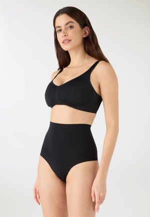 Donna che indossa un reggiseno nero senza cuciture e mutande a vita alta, in piedi contro uno sfondo chiaro uniforme, leggermente rivolta verso destra.