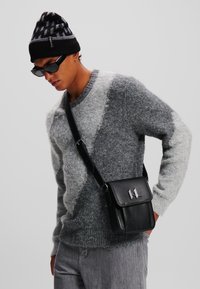 Maglione grigio e bianco con motivo, borsa a tracolla in pelle nera con hardware argentato, jeans scuri, berretto nero con design geometrico, occhiali da sole neri.