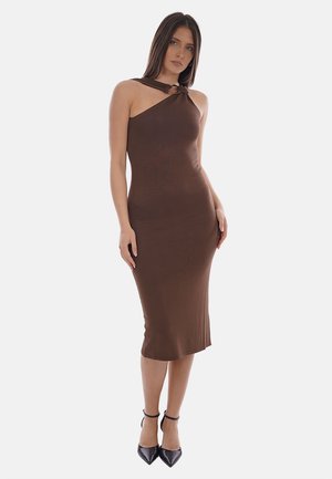 Donna in piedi che indossa un abito midi marrone aderente senza maniche con scollo halter annodato e tacchi neri con cinturino alla caviglia su sfondo bianco.