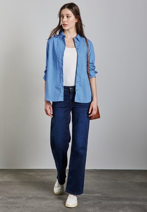 Blauw gestreepte overhemddag over een witte tanktop, gecombineerd met donkerblauwe wijde jeans en witte sneakers, terwijl hij/zij een bruine schoudertas vasthoudt.