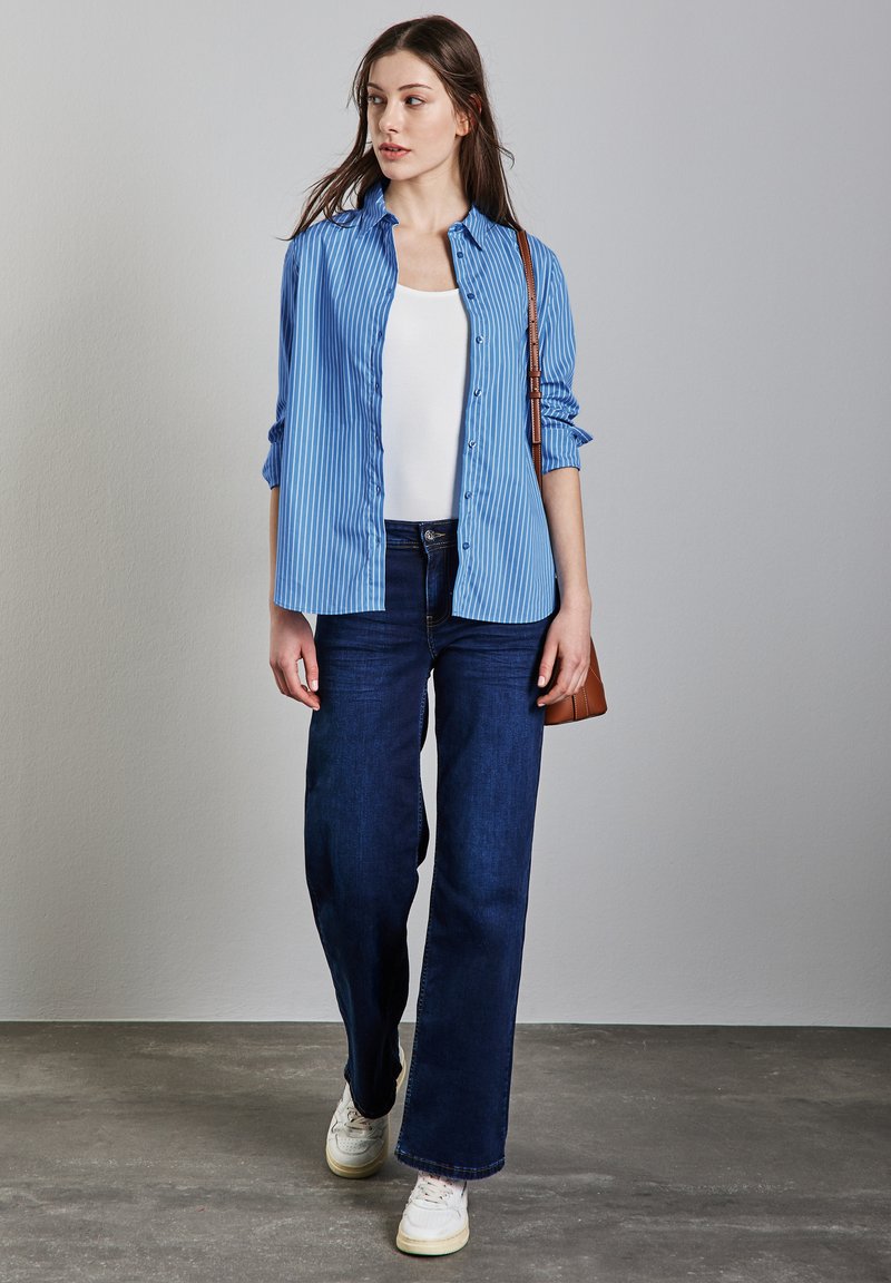 Camicia a righe blu con bottoni indossata sopra una canotta bianca, abbinata a jeans a gamba larga blu scuro e sneaker bianche, mentre tiene una borsa a tracolla marrone.
