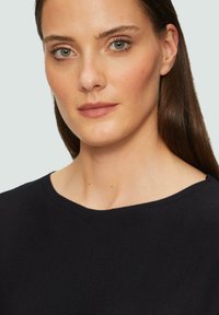 Un pull navy foncé avec un large décolleté, une texture côtelée et un tissu lisse, porté par un mannequin avec des cheveux bruns raides et un maquillage neutre.