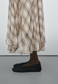 Brauner karierter, transparenter, geschichteter Rock mit Rüschensaum und schwarzen Fell-Pantoffeln. Die Schuhe zeichnen sich durch eine verlängerte Form und weiche Textur aus.