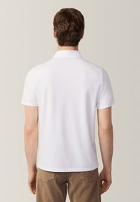 Polo shirt bianco a maniche corte con colletto, texture liscia e orlo dritto; indossato con pantaloni khaki, visto da dietro.