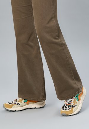 Braune Hose kombiniert mit mehrfarbigen Sneakers mit beigen, orangen, grünen und schwarzen Leopardenmuster-Details auf grauem Bodenhintergrund.