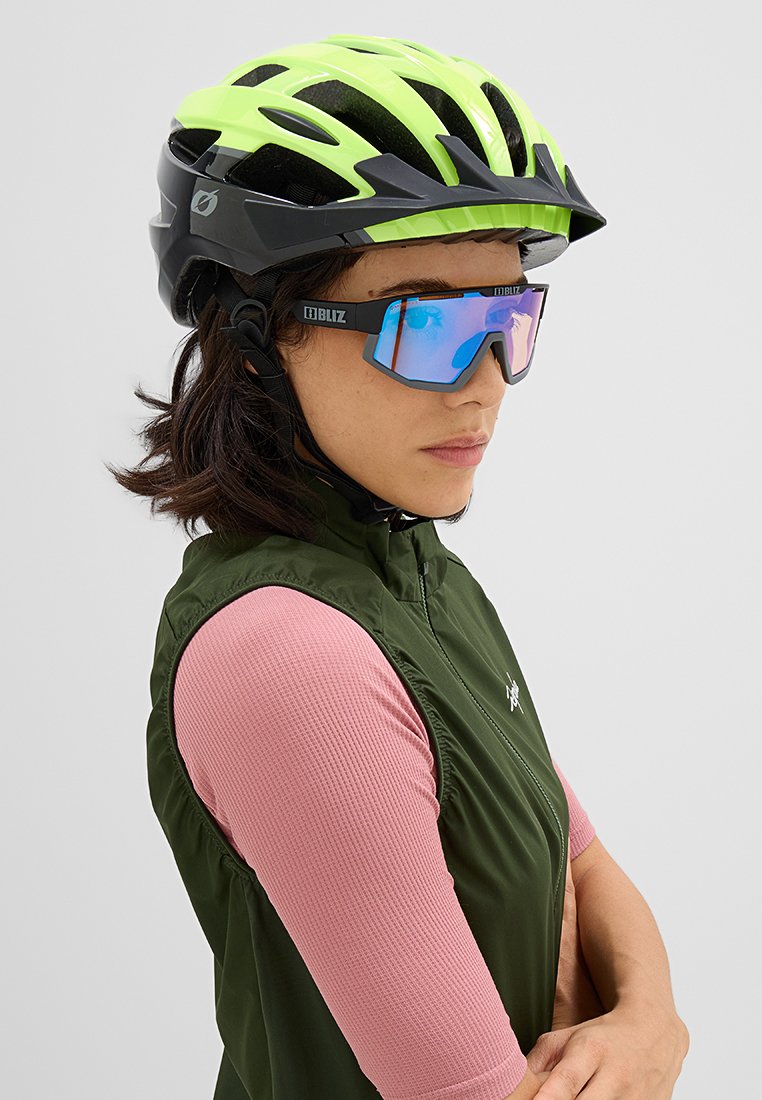 Leichter schwarzer Fahrradhelm mit neon-gelben Akzenten und Belüftungslöchern. Das Modell trägt eine verspiegelte Sonnenbrille sowie ein sportliches Oberteil in Pink und Grün.