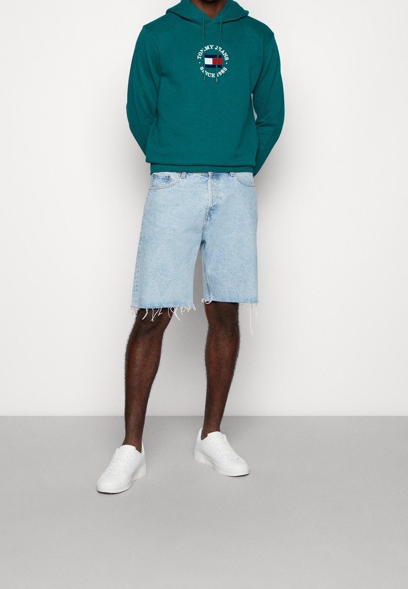 Homme portant un sweat à capuche teal de Tommy Jeans, un short en denim bleu clair effiloché et des baskets blanches, debout devant un fond blanc uni.