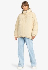 Giacca imbottita beige con cappuccio, due tasche frontali con zip, polsini elasticizzati e orlo regolabile con cordino. Abbinata a jeans a gamba larga blu chiaro.