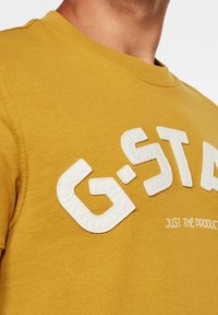 Mosterdgele t-shirt met gestructureerde witte letters die "G-STAR" spellen en "JUST THE PRODUCT" eronder. Ronde hals, korte mouwen.