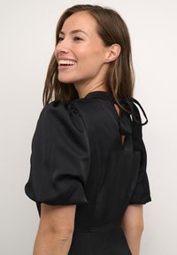 Rochie din satin negru cu mâneci bufante scurte, detaliu cu decupaj la spate și accent legat. Textură netedă și croială ajustată.