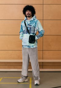 Το φούτερ διαθέτει σχέδιο καμουφλάζ σε μπλε-πράσινο χρώμα με μια μπροστινή τσέπη. Το παντελόνι είναι γκρι κορντόρι με πλευρικές ρίγες. Το αξεσουάρ είναι μια μικρή μαύρη τσάντα.