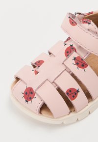 Rosa barnesandaler med røde marihøneprint, åpen tå, justerbar stropp og hvit såle på hvit bakgrunn.