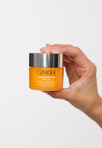 Orange burk med Clinique Superdefense SPF 40 gel, med ett silverfärgat metallock och vit text som visar produktinformation. Hand som håller burken.