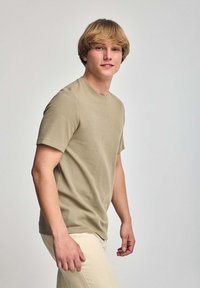 Korte mouwen, ronde hals t-shirt in licht olijfgroen, gemaakt van textuurstof. Model draagt een lichte broek en staat op een neutrale achtergrond.