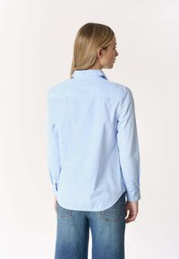 Donna con capelli biondi lisci, che indossa una camicia a righe azzurra a bottoni e jeans blu, vista di schiena contro uno sfondo semplice.