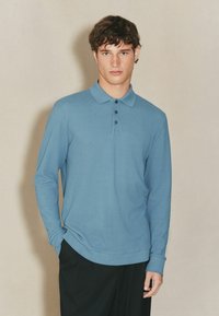 Polo a maniche lunghe in tessuto leggero blu chiaro con colletto, placca con tre bottoni e vestibilità rilassata. Indossato con pantaloni scuri.