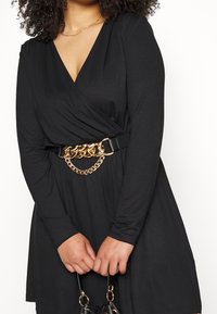 Femme portant une robe portefeuille noire à manches longues avec une ceinture chaîne dorée, tenant un sac à main noir avec des ornements dorés devant un fond blanc.