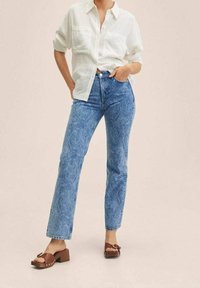 Jeans de color azul claro con estampado de paisley, de cintura alta y corte recto. Combinados con una camisa blanca de botones y sandalias marrones de tacón cuadrado.
