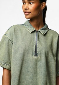 Prevelika zelena traper košulja s reverom na patentni zatvarač, kratkim rukavima i mekim teksturama. Ima suptilan izlizani uzorak.