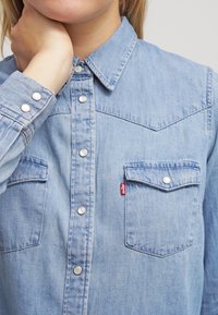 Kvinna som bär en ljusblå denimskjorta med tryckknappar och bröstfickor, hand som rör vid nacken, inget synligt ansikte.