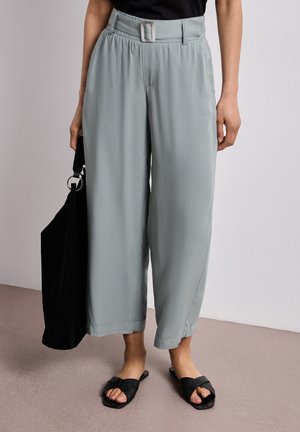 Personne portant un pantalon gris clair taille haute à jambes larges avec une ceinture, un haut noir, des sandales noires, et tenant un grand sac cabas noir.