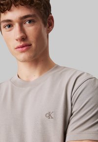 Szara bawełniana koszulka z okrągłym dekoltem i krótkimi rękawami, z haftowanym logo "CK" na lewej piersi. Gładka tekstura.