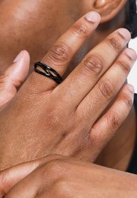 Anello a spirale nero indossato su una mano con pelle strutturata. L'anello presenta un design a più fili, che contrasta con il tono della pelle.