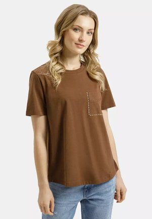 Kobieta o falowanych blond włosach, ubrana w brązowy t-shirt z krótkim rękawem i kieszenią ozdobioną ćwiekami oraz jasnoniebieskie dżinsy, stojąca na jednolitym tle.