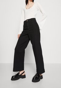 Žena v bílém strukturovaném dlouhém triku s dlouhým rukávem, vysokých širokých černých kalhotách s vysokým pasem a černých platformových pantoflích na podpatku se sponami.