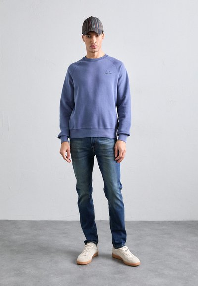 Blå bomullsgenser med rund hals og ribbestrikkede mansjetter, sammen med mørkeblå jeans og beige sneakers. Stripete caps fullfører looket.