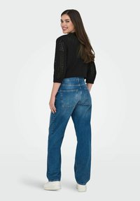 Crna pletena majica s tri četvrtine rukava, uparena s trapericama viskog struka u plavoj boji i bijelim tenisicama. Glatka tekstura i opušteni kroj.