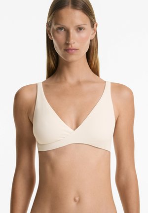 Bralette beige côtelé avec un décolleté en V profond, des bretelles larges et un panneau avant superposé. Le tissu semble doux avec de subtiles détails texturés.
