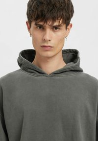 Šedá mikina s kapucí z měkké, texturované tkaniny a volným střihem. Kapuce je přiléhavá s viditelnými šicími detaily.