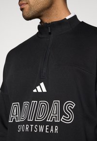 Μαύρο φούτερ με φερμουάρ, κατασκευασμένο από απαλό ύφασμα, με ψηλό γιακά, λευκό λογότυπο "ADIDAS" και σχέδιο με τρεις λευκές ρίγες στο στήθος.