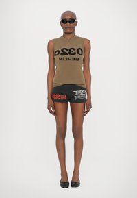 Top senza maniche color oliva con testo nero "0320 BERLINO", abbinato a pantaloni corti neri con grafiche rosse. Il modello indossa occhiali da sole neri e ballerine.