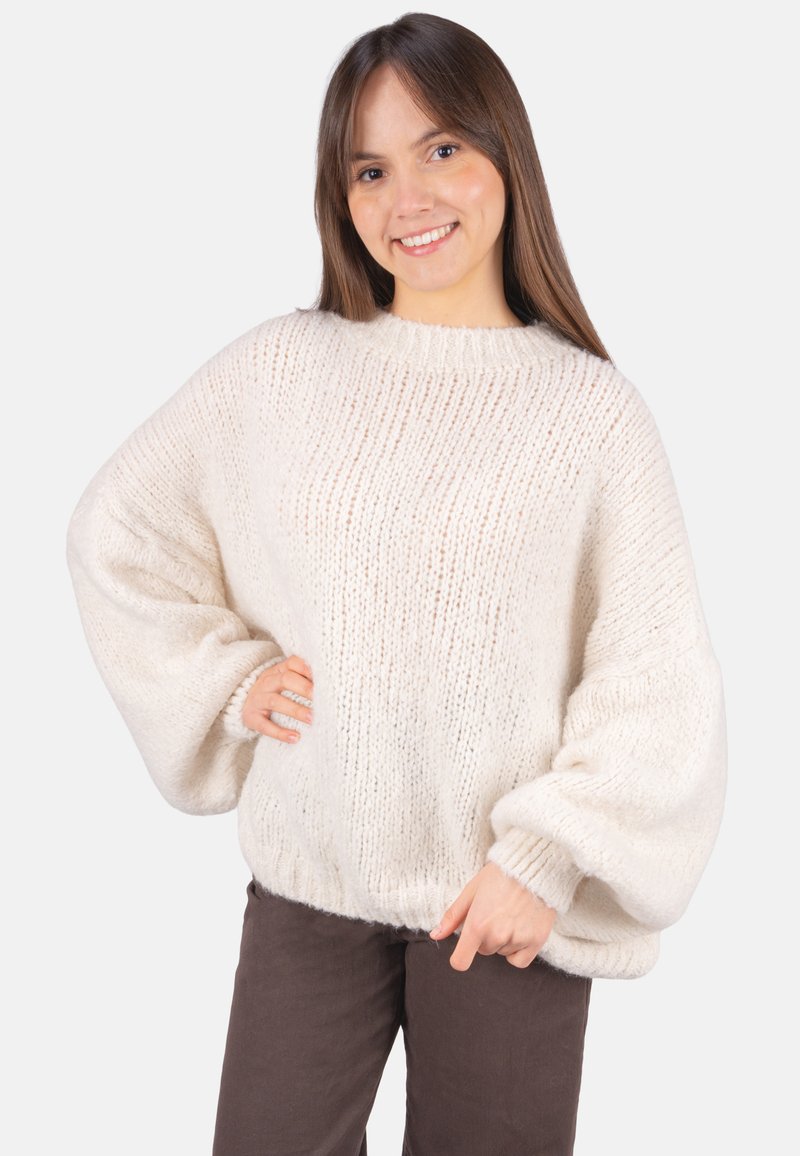 Cremefarbener Pullover mit lockerem, übergroßem Design, gerippter Struktur und breiten Ärmeln. Wird über dunkelbraunen Hosen getragen, mit rundem Ausschnitt.