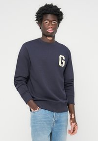 Marineblå hettegenser med ribbet rund hals og G-logo-lap på venstre bryst, kombinert med lyseblå jeans. Jevn tekstur, avslappet passform.