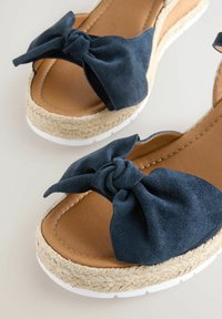 Sandali in suede blu navy con un grande fiocco sulla parte superiore, una soletta in pelle color tan e una suola in juta testurizzata con bordi in gomma bianca.