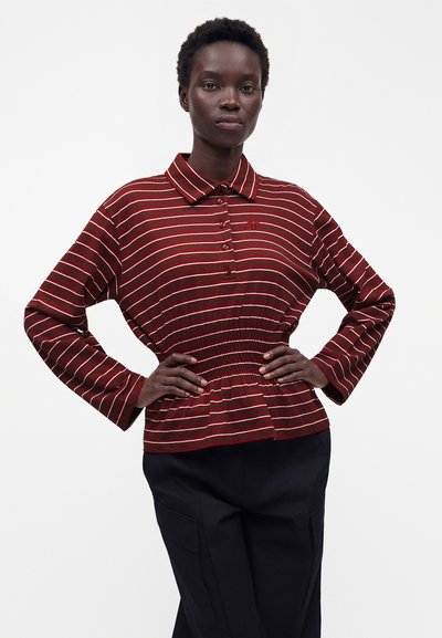 Femme aux cheveux courts et bouclés portant un haut rouge et blanc rayé à manches longues avec une taille froncée, debout les mains sur les hanches.