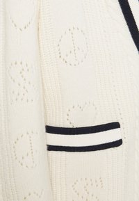 Tissu tricot crème avec des signes de paix perforés et des cœurs, avec une bordure rayée noire et blanche.