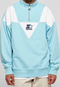 Ljusblå sweatshirt med en vit triangulär design, halvdragkedja vid kragen och broderad STAR-logotyp på bröstet. Mjuk tyg, ribbade manschetter.
