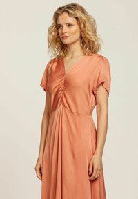 Persikofärgad klänning med korta ärmar, V-formad halsringning, insamling i mitten, flödande kjol. Slät tyg med en mjuk lyster.