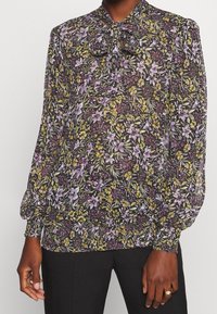 Durchsichtige Bluse mit floralem Muster auf dunklem Hintergrund in Lila-, Gelb- und Grüntönen. Sie verfügt über einen Schleifen-Ausschnitt sowie elastische Bündchen und einen elastischen Saum.